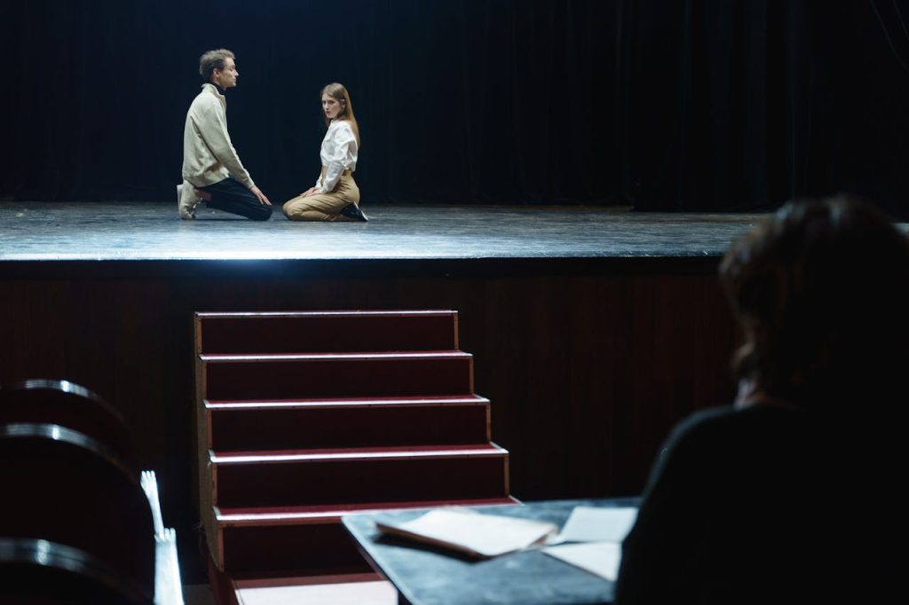 Actors in a dramatic rehearsal on stage, under dim theater lights.