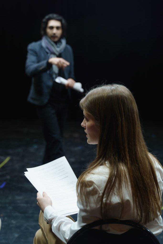 Actors rehearse a dramatic scene in a dimly lit theater room, focusing on dialogue and performance.